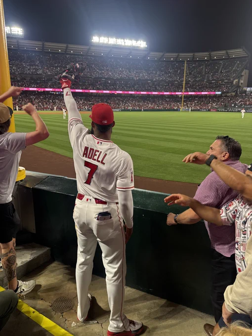 A photo of Jo Adell, his back towards the camera, standing in the right field stands and holding up his glove to show off the third home run robbery catch of the evening at Angel Stadium.