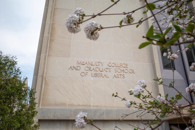 Exterior stone facade of McAnulty College and Graduate School of Liberal Arts building, with bronze lettering visible through foreground branches of white spring blossoms.