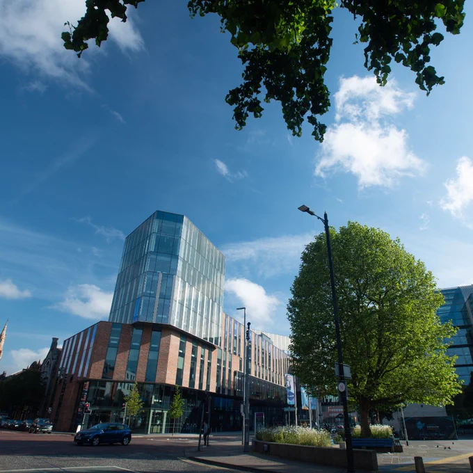 Modern glass and brick Ulster University Belfast campus building under a bright blue sky with scattered clouds and leafy trees in the foreground.