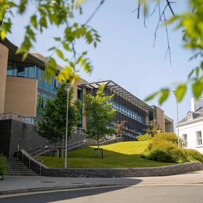 Modern Derry~Londonderry campus building with large glass windows and a sloped green lawn under a clear blue sky.