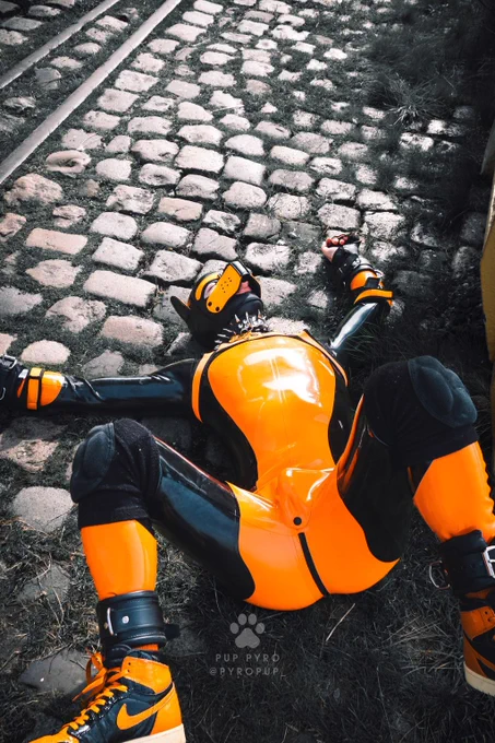 GimpSimp lying on a cobbled surface in orange pup suit, pup hood, harness and cuffs