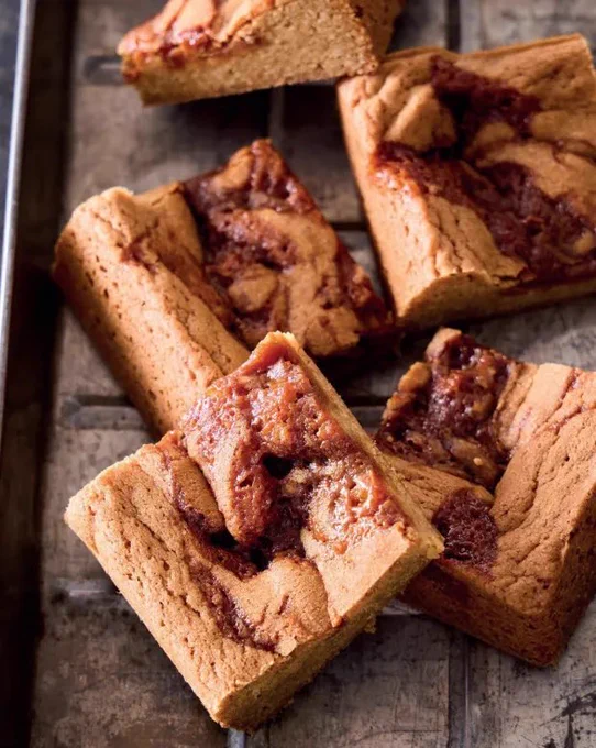 Golden brown blondies cut into thick bars with a glossy caramel swirl running through the center, showing a dense, chewy texture and slightly crackled top, arranged on a rustic wooden surface with a rich, buttery finish.