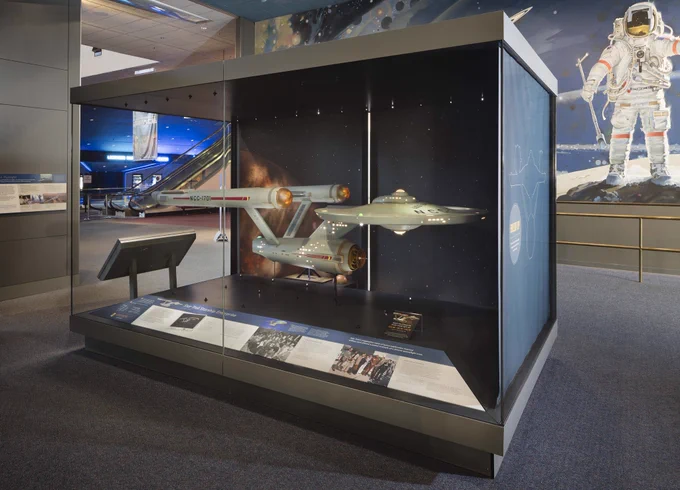 Exhibit at the Smithsonian National Air and Space Museum featuring a display of Star Trek starships, including the famous USS Enterprise model, and a space suit. Informational panels are visible in the foreground.