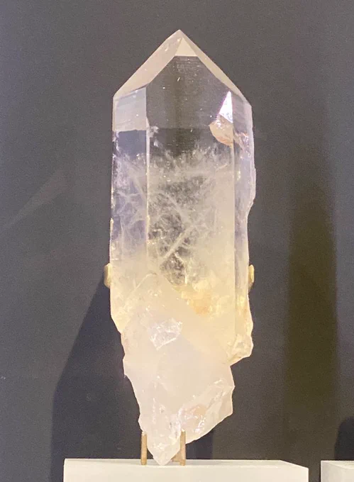 Long vertical quartz crystal with a transparent pyramidal top. The bottom is milkier with jagged edges. It sits on a metal mount on a frosted plinth in front of a grey background. At the Royal Ontario Museum.