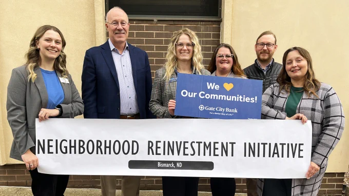 Group of people holding a banner that reads ‘Neighborhood Reinvestment Initiative – Bismarck, ND,’ with additional people holding a sign that says ‘We ♥ Our Communities! Gate City Bank’ in front of a brick building.