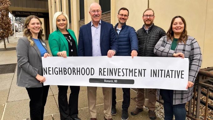 Group of people standing on a city sidewalk holding a banner that reads ‘Neighborhood Reinvestment Initiative – Bismarck, ND’ in front of the City/County Office Building.