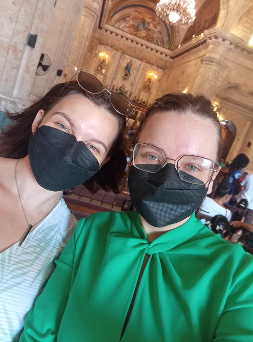 Two women in black boat style KN95 masks smile in front of the Virgin Mary