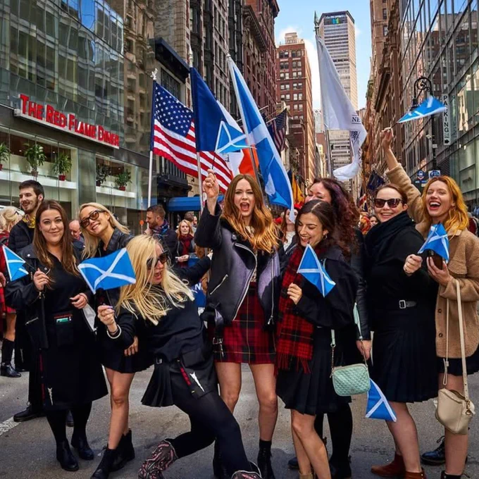 Group of people waving Scottish flags during a street parade in a city with tall buildings.