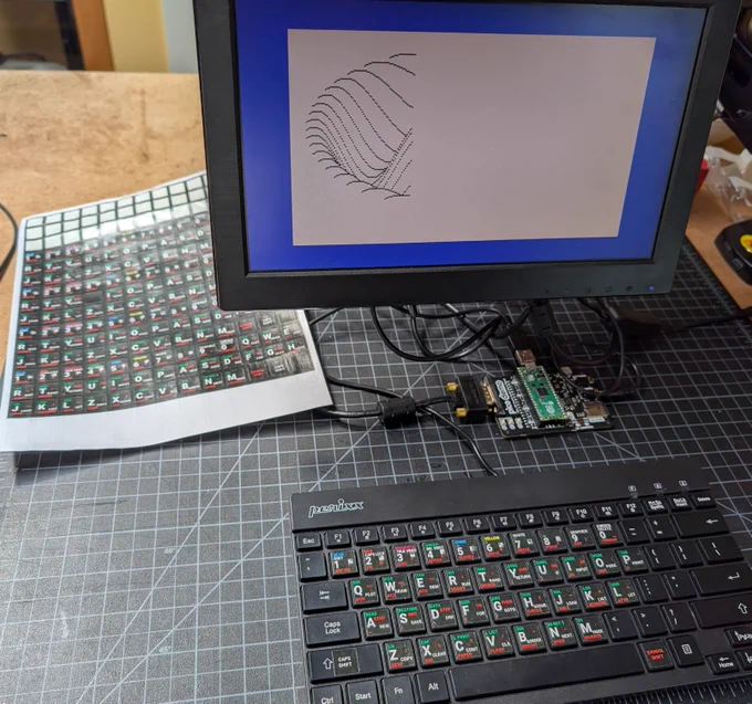 A photo of a USB keyboard, a Raspberry Pi Pico mounted on a PCB, a computer monitor, and a partially-used sheet of stickers. Some of the stickers have been applied to the keyboard. The stickers replicate the special keywords and characters that were silkscreened on the original ZX Spectrum keyboard.