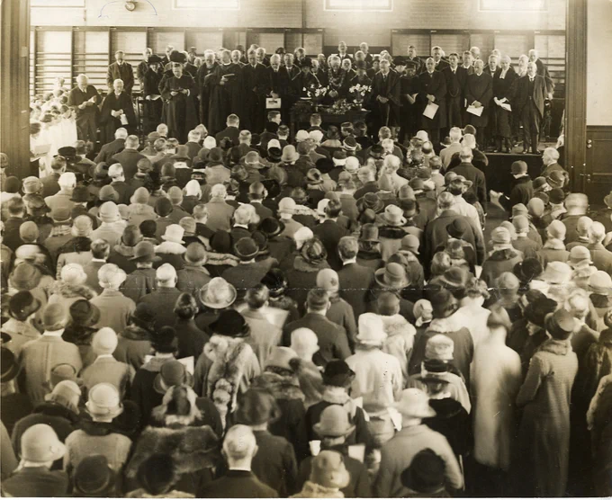 A group of people stand on a stage facing an even larger group, whose backs are to the camera. A black and white photo.