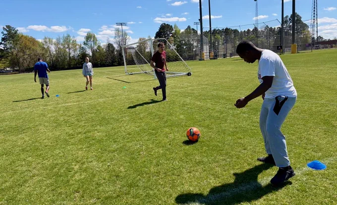 Four people are on a grassy soccer field, two near a goal post and one dribbling an orange and black soccer ball. The scene is set under a clear blue sky with trees in the background.
