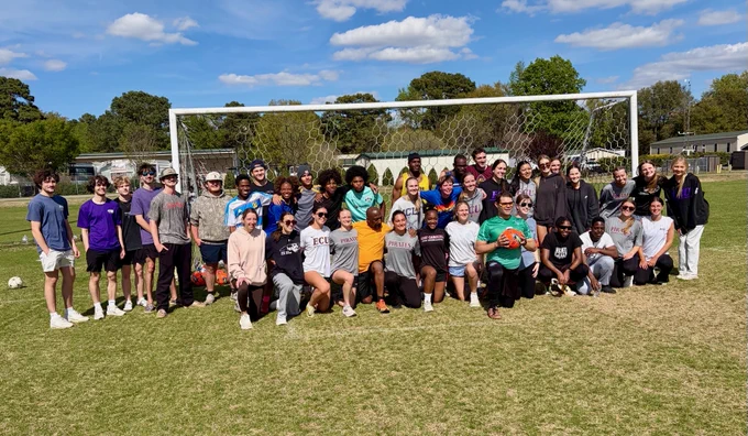 A large group of people posing in front of a soccer goal on a grassy field. Some individuals are holding sports equipment like a soccer ball. The setting is outdoors with trees and blue sky in the background.