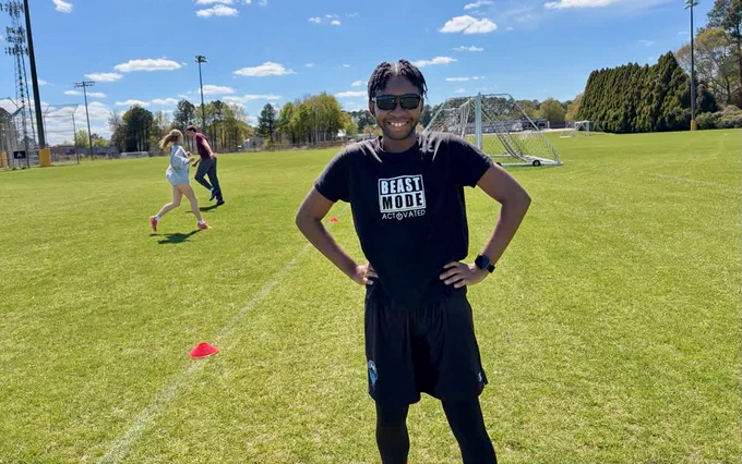 A person stands smiling on a grassy sports field wearing sunglasses and a black t‑shirt with "Beast Mode Activated" printed on it. Two other people are in the background near cones. The sky is clear and sunny.