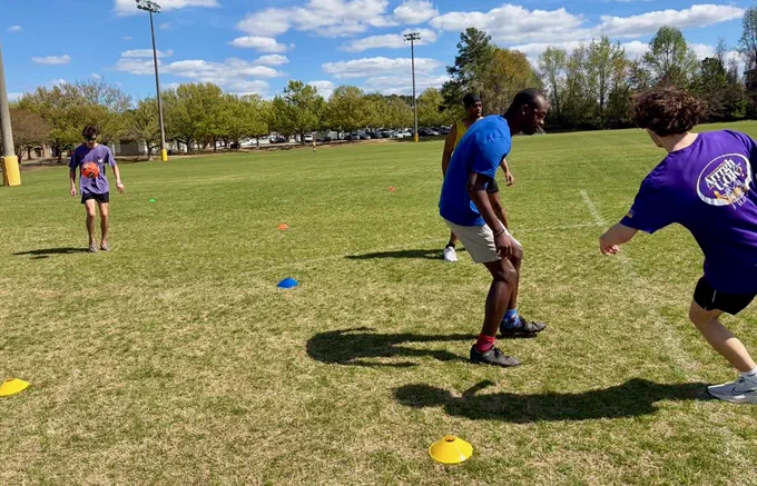 Four people playing soccer on a grassy field with colorful cones marking boundaries. One person is about to kick the ball while others are positioned on the field. 