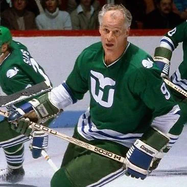 Gordie Howe skating on the ice during a game, wearing a green Hartford Whalers uniform with the team’s whale-tail logo on the chest. He is holding a hockey stick in both hands and leaning slightly forward in motion. He has short, thinning gray hair and is not wearing a helmet. Teammates in similar green uniforms are visible behind him, and spectators can be seen seated in the arena stands in the background.
