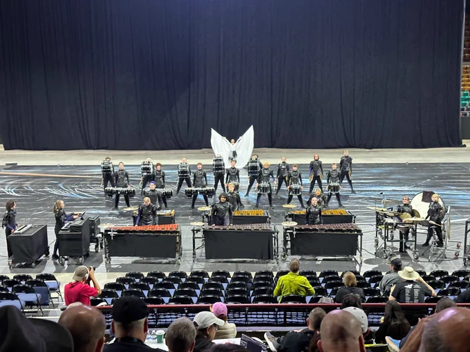 An indoor percussion ensemble performs on a gymnasium or arena floor. Approximately 25 young performers dressed in black sparkle uniforms are arranged in a semicircle formation. A central performer dressed in white holds large white fabric wings outstretched above their head. In the foreground, several performers stand at marimba and xylophone keyboard percussion instruments, while rows of snare drummers stand behind them. The performance floor is dark with abstract line designs. Audience members are visible in the foreground seats.