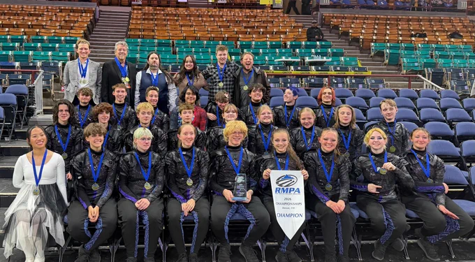 A large group of approximately 30 young musicians, directors, and support staff pose for a group photo in the stands of an arena in Denver, Colorado. Most performers wear black sparkle uniforms with purple accents and blue-ribboned gold medals around their necks. One performer on the far left wears a white costume. Two members in the front row hold a white banner reading "RMPA Championships, Denver, CO — CHAMPION" and a 1st Place trophy award. Adults in street clothes, also wearing medals, are visible in the back rows.