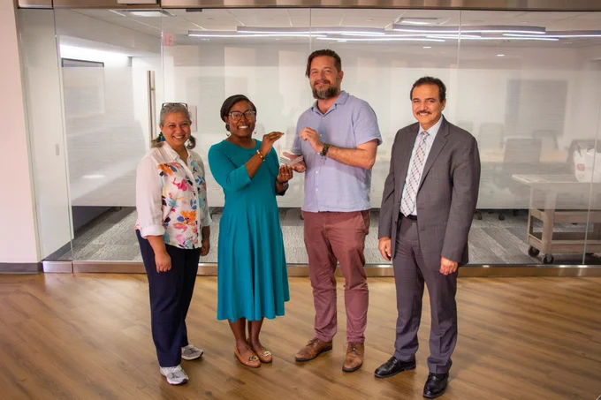 Dr. Sallam, Dr. Hill, Dr. Goodman, and Dr. Serag stand together in front of a glass-walled conference room as Dr. Hill presents the inaugural PHHD Pilot Award to Dr. Goodman. Dr. Goodman holds the clear award at center while the group smiles for the camera.
