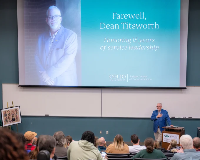 A green screen reads, "Farewell, Dean Titsworth" with "Honoring 15 years of service leadership" under it, with a portrait of Dean Scott Titsworth, with the Dean standing at a podium below it, with his hand placed on his heart.