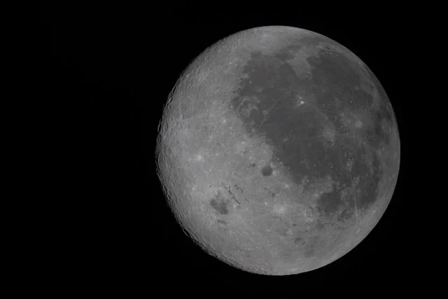The Moon against the black of space. The right half is heavily marked with darker splotches and craters, including a very large patch that fills most of the upper right quadrant. The left side of the Moon, which is not visible from Earth, is much more uniformly gray. It has small divots and craters, but they do not change the surface color. There is one prominent darker spot between 7 and 8 o'clock on the Moon's surface. 