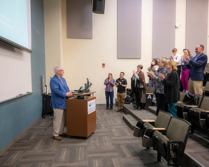Dean Scott Titsworth stands at the podium, with attendees standing to clap, including Ohio University President Lori Gonzalez.