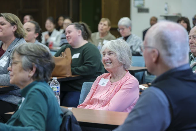 A classroom setting shows a diverse group of adults seated at desks, listening attentively. Mary Andrew is in focus, smiling with a name tag visible. The scene suggests a lecture or workshop environment.