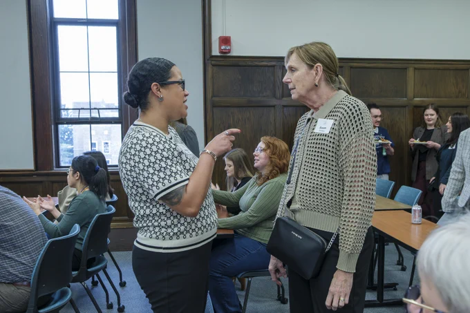 In a warmly lit room, two women converse while others sit at tables or stand eating. One woman wears a \"Susan text@1\" name tag. The setting features wood paneling and a window providing natural light, enhancing the collegiate atmosphere.