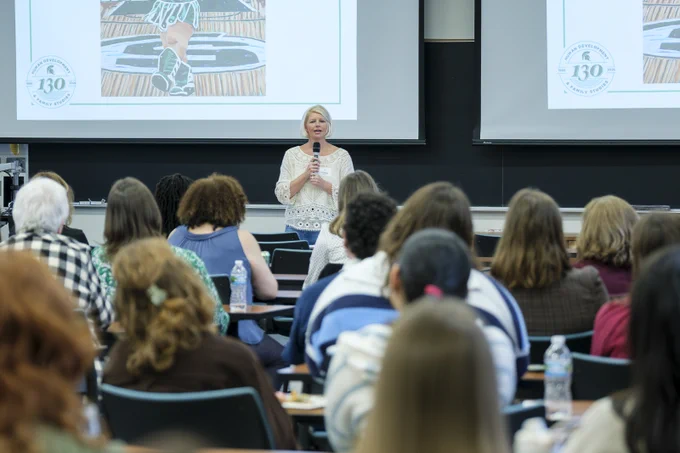 A fair-skinned, blonde-haired woman stands speaking into a microphone to a crowd of seated people. Two projector screens display \"HUMAN DEVELOPMENT@130 & FAMILY STUDIES\". The speaker wears a white shirt and jeans.