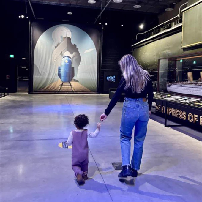 Une femme et un enfant, main dans la main, se dirigent vers une grande maquette de train illuminée dans la salle d'un musée.

