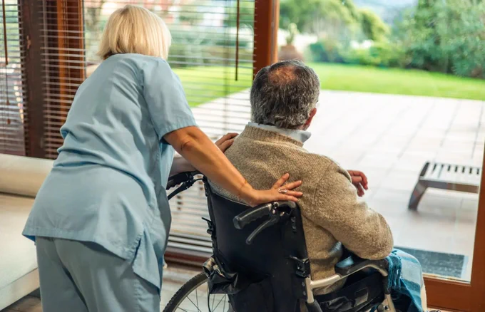 An individual in a wheelchair is positioned near large windows, overlooking a garden, with another person standing beside and gently holding the wheelchair.
