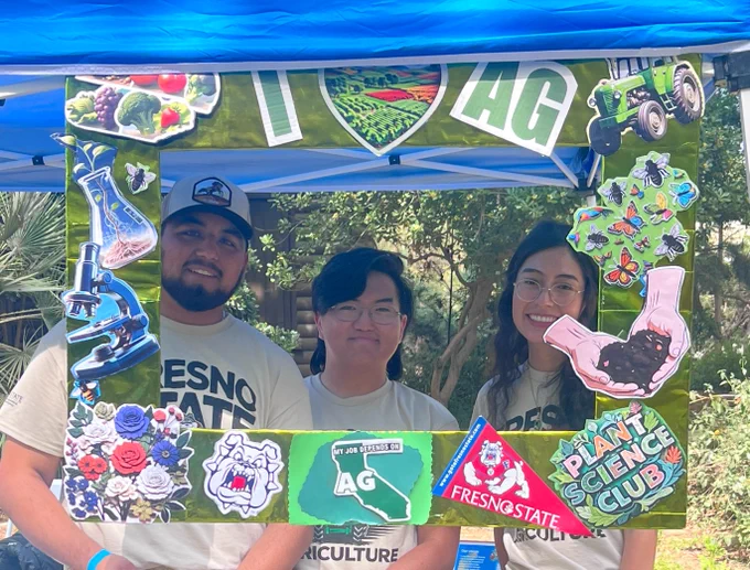 Three people stand together smiling under a blue canopy. They hold a decorative frame with agricultural-themed images, including lab equipment, a tractor, and plants. The frame has labels like "Fresno State," "Plant Science Club," and "I ❤️ AG." In the background, there are trees and greenery.