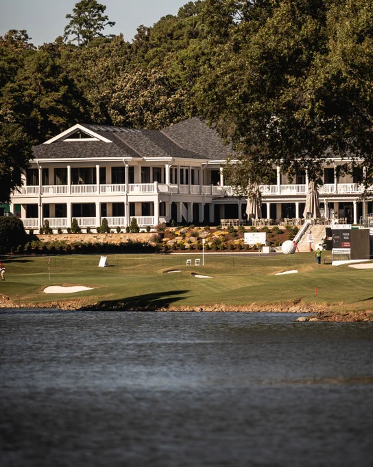 A large, elegant clubhouse with multiple white verandas is surrounded by lush greenery and trees. It overlooks a well-manicured golf course with sand traps, and there’s a body of water in the foreground.