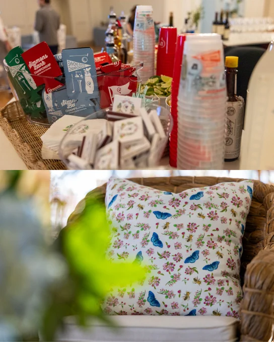 The image shows a countertop with an assortment of bar items, including cocktail mixers, branded koozies and stacked plastic cups. In the background, various beverage bottles are visible. Below, a wicker chair is adorned with a cushion featuring blue butterflies and pink flowers. A blurred plant is in the foreground.