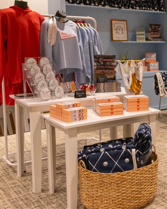 A display in a store featuring University of Pennsylvania merchandise, including apparel, books, and accessories. A basket with blankets is on the floor beside the tables.