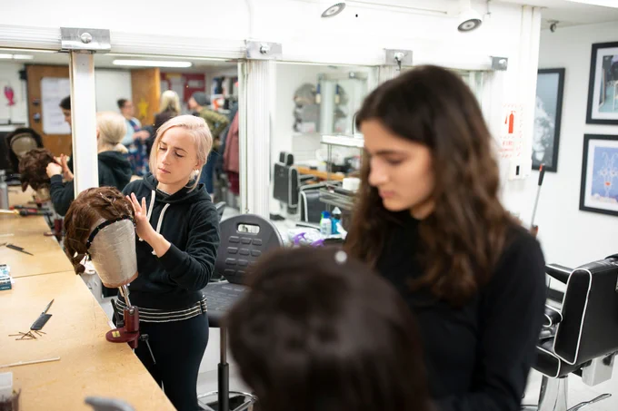 A group of individuals in a makeup and hairstyling studio are working with wigs on mannequin heads. The room is filled with styling equipment, mirrors, and chairs.