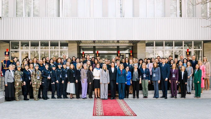 A large group of people are gathered in front of a building for a formal event. Many are wearing uniforms or business attire. A red carpet is laid out in front of them.