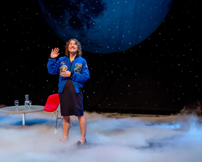 Christina Koch waves to the audience on a stage, wearing a blue NASA jacket.