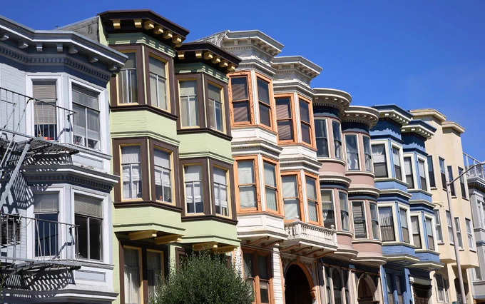 Colorful row of Victorian-style houses with ornate bay windows and decorative trim, painted in pastel shades like blue, green, cream, and peach, under a clear blue sky.