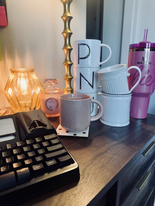 Freewrite typewriter, some candles and a slew of stacked coffee mugs on a brown desk.