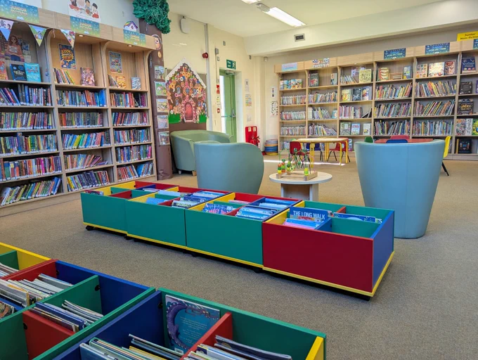A colorrful children's library features shelves filled with books, cozy armchairs, and low tables. Bright bins contain more books, creating an inviting and playful atmosphere.