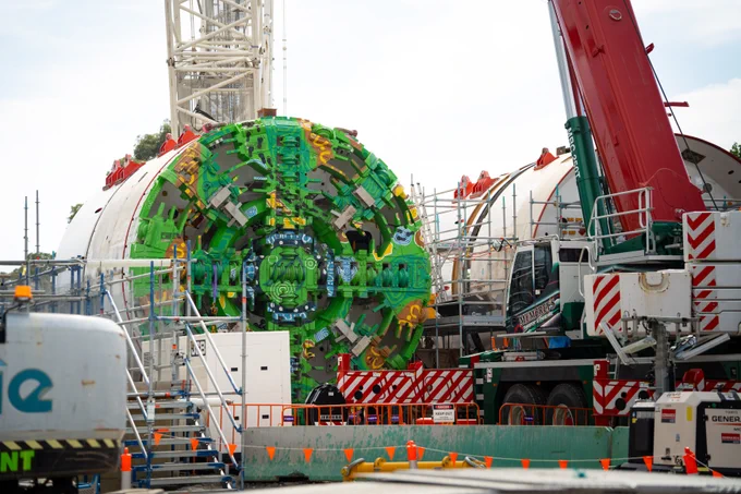 A large tunnel boring machine with a vibrant green front is under assembly at a construction site. The machinery is surrounded by cranes and various construction vehicles, including a prominent red crane. Orange safety cones and barriers are scattered around the site, ensuring a secure perimeter.