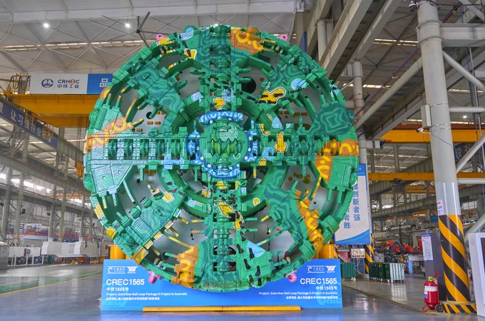 A large green tunnel boring machine is inside an industrial warehouse. The machine is surrounded by a spacious interior with structural beams.