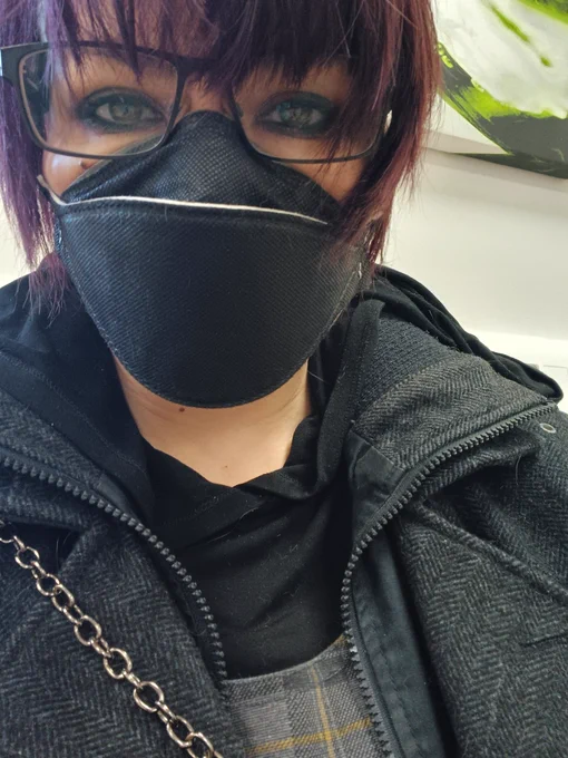 Humanoid in black respirator mask and glasses, with short purplish hair, in black and grey clothing. 