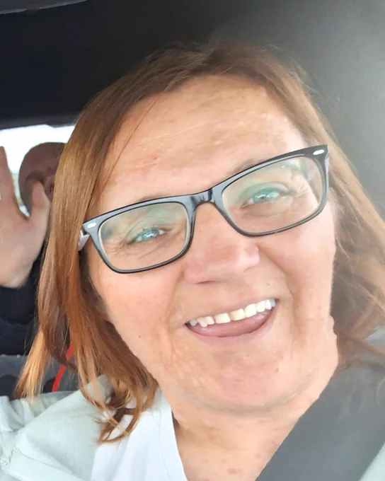 Hayley, a white woman with shoulder-length light brunette hair, sits in a car and takes a selfie. She looks animated and smiley. Her NF1 presents as small bumps on her face.
