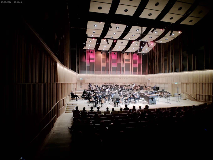 A wide-angle view from the back of a concert hall during the SFE Spring Concerts 2026. A large youth orchestra is performing on a brightly lit, modern wood-paneled stage under an acoustic ceiling. An audience is seated in the foreground, watching the performance, which includes a variety of instruments and percussion.