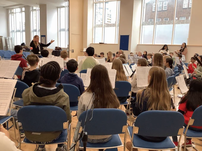 A large group of young musicians seated in a bright rehearsal hall during Great Big Woodwind Day 2026. Viewed from behind, the students are focused on their sheet music while a female conductor at the front of the room leads the ensemble with expressive hand gestures.
