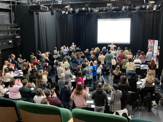 A wide shot of music educators and professionals gathered for a presentation at the Birmingham Music Education Conference, hosted by Services For Education. The image captures the scale of the active workshop taking place in a theatre-style setting.