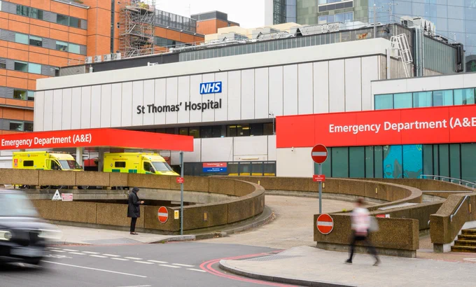 Outdoor view of St Thomas' Hospital Emergency Department. Includes the name of the hospital on a grey building, with red signage in front saying Emergency Department (A&E). There are two ambulances in front.