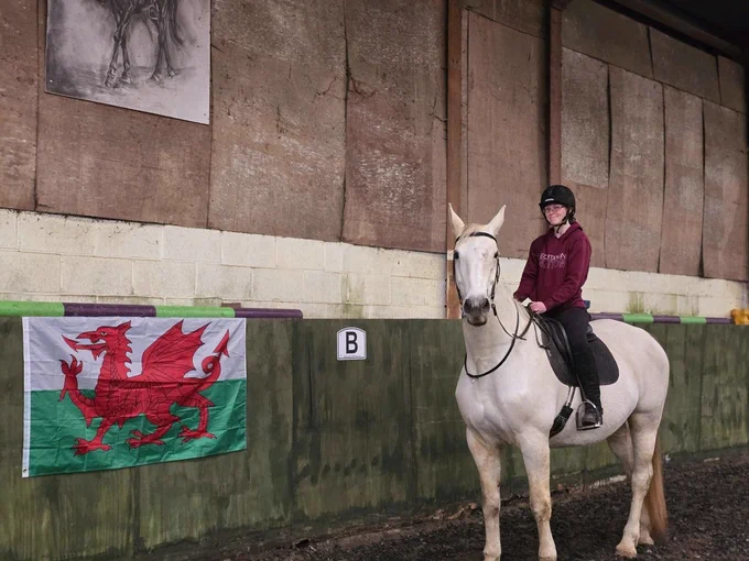 A white horse looks at the camera as it stands before a Welsh flag. The rider is smiling.