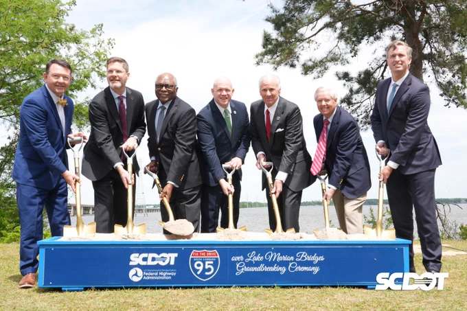 A group of seven individuals in suits is participating in a groundbreaking ceremony at the Lake Marion Bridge. They are holding shovels with sand, standing behind a sign that includes the SCDOT and I-95 logos. Trees and water are visible in the background.